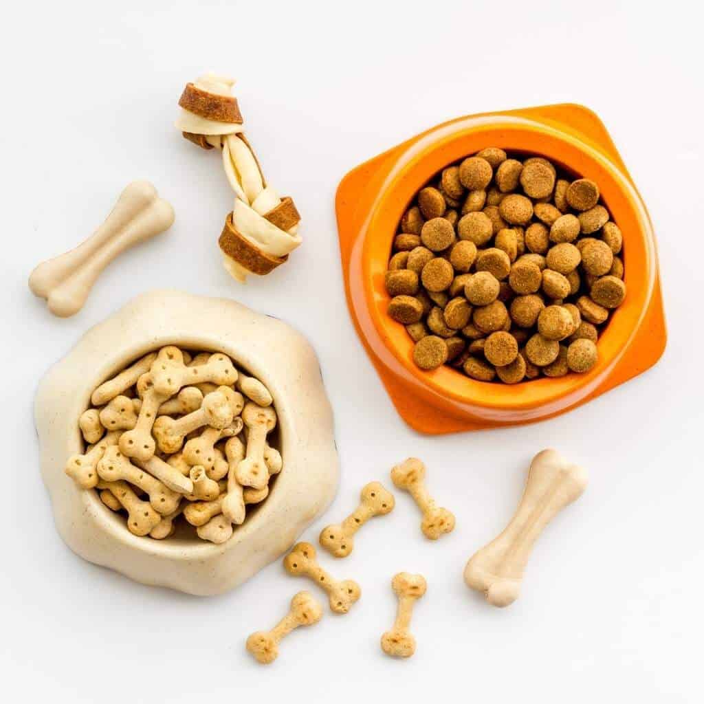a bowl of dog food, a bowl of dog biscuits in the shape of bones, next to other dog treats