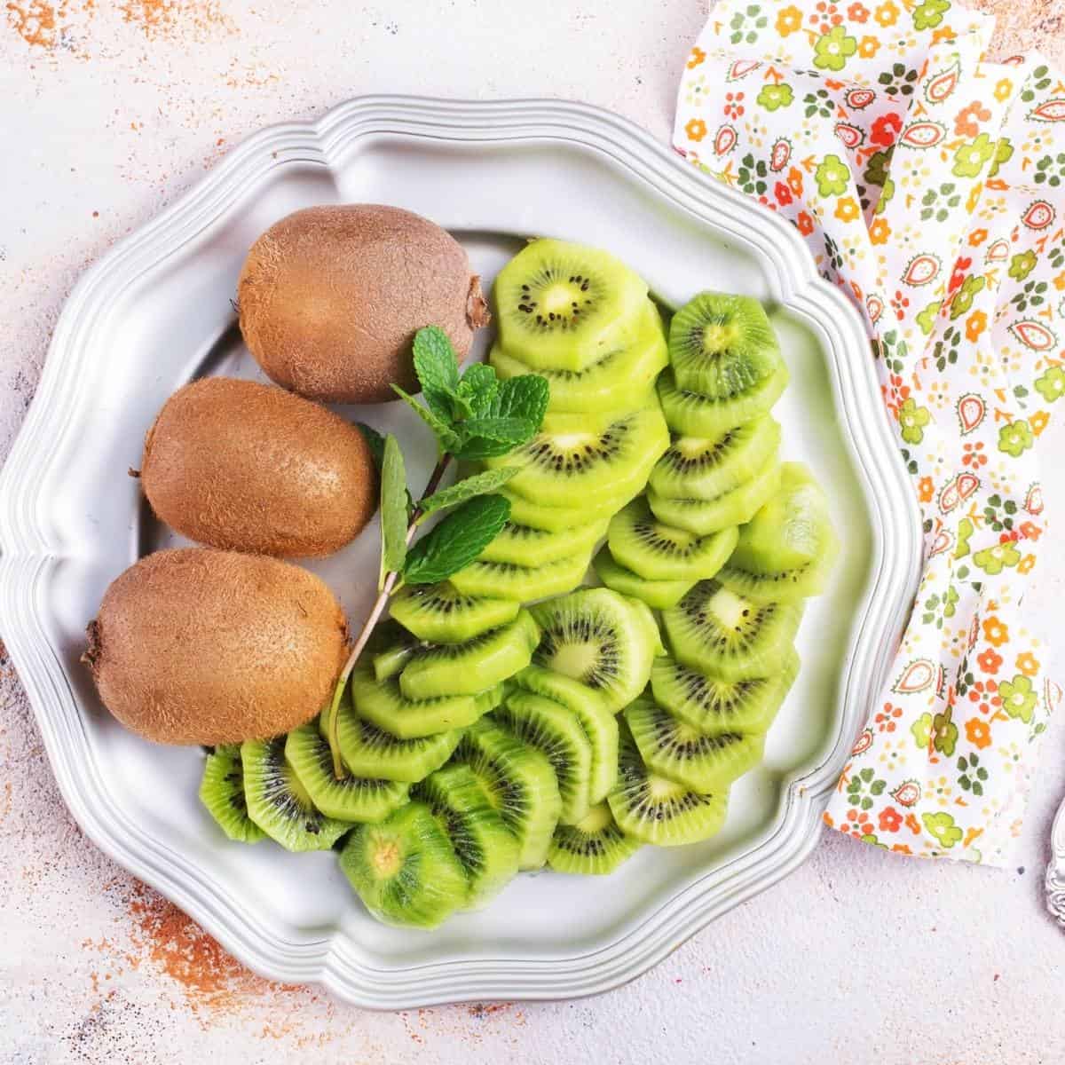 slices of kiwi and whole kiwi fruits on a white plate next to a flower dish towel